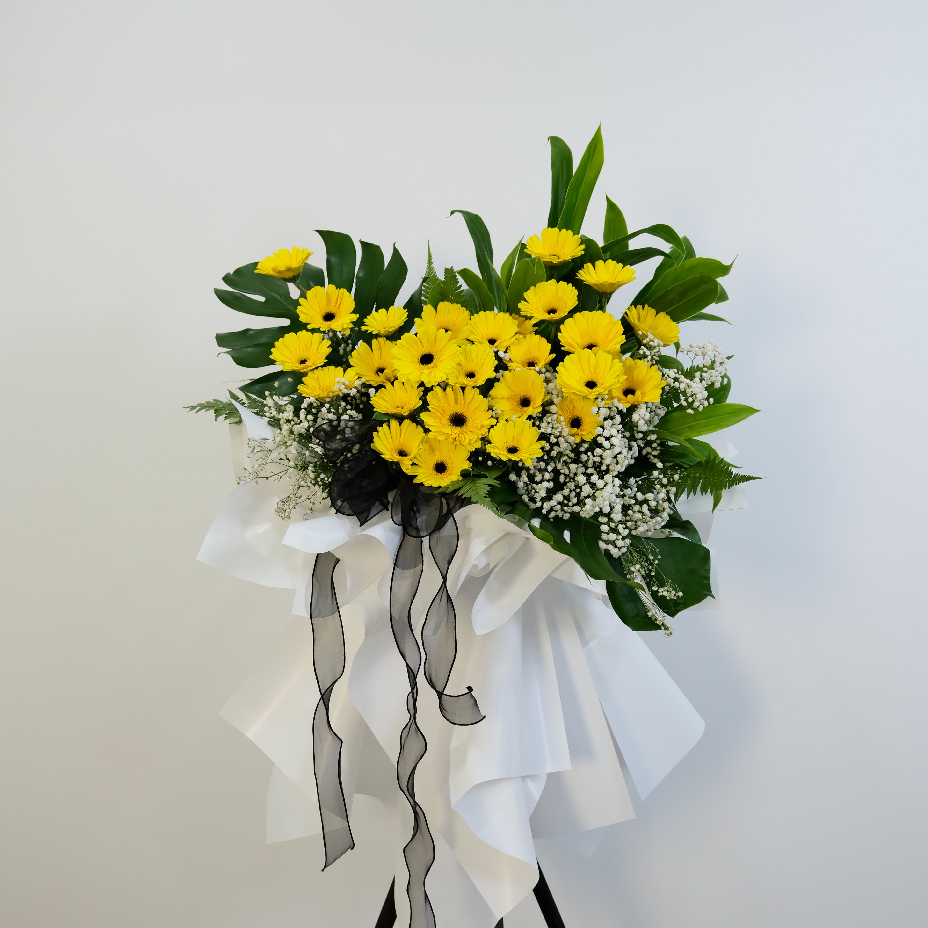 condolence flower stand of yellow flowers with green leaves and ribbons on a light gray background