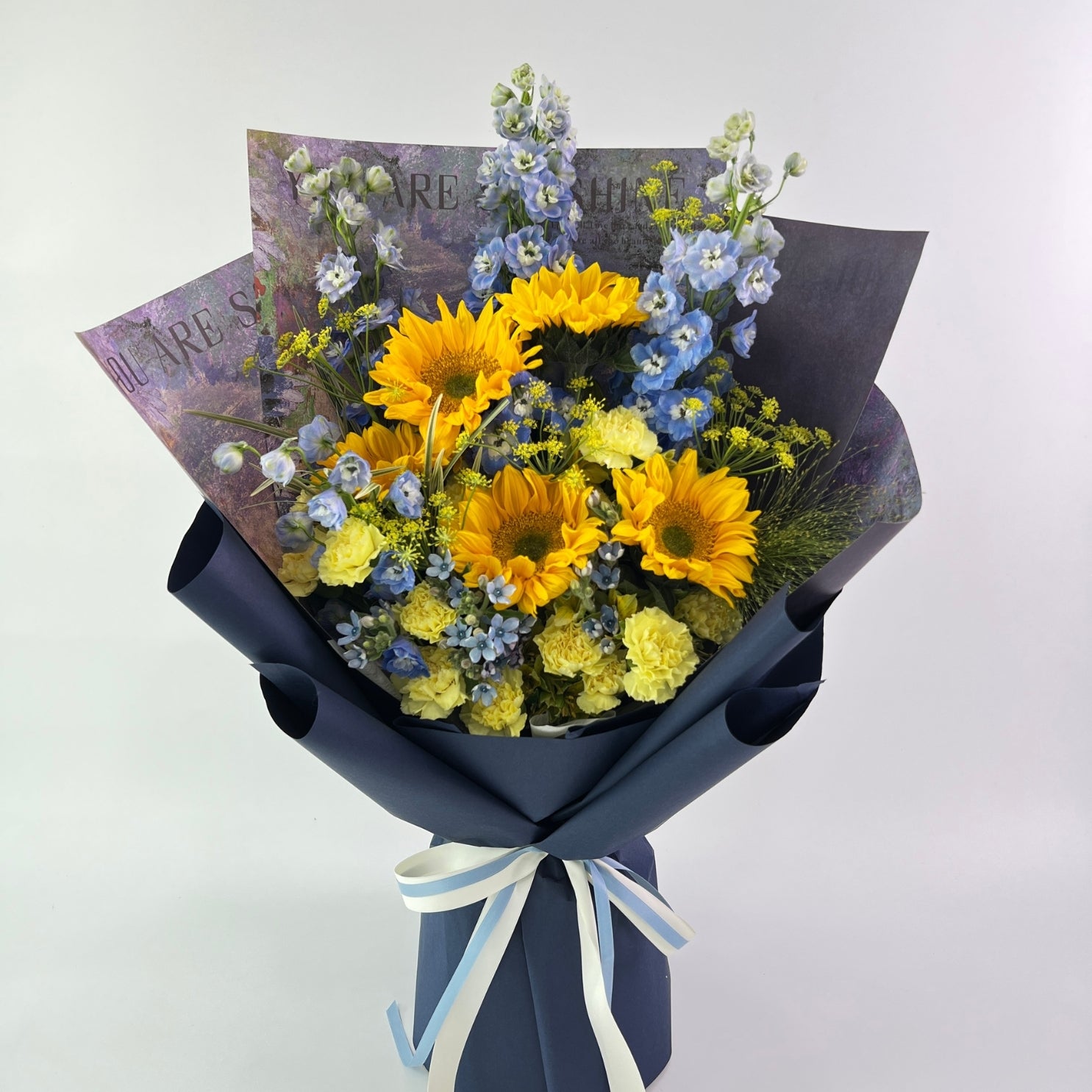 Bouquet of yellow and blue flowers wrapped in black paper with a white ribbon on a light gray background.