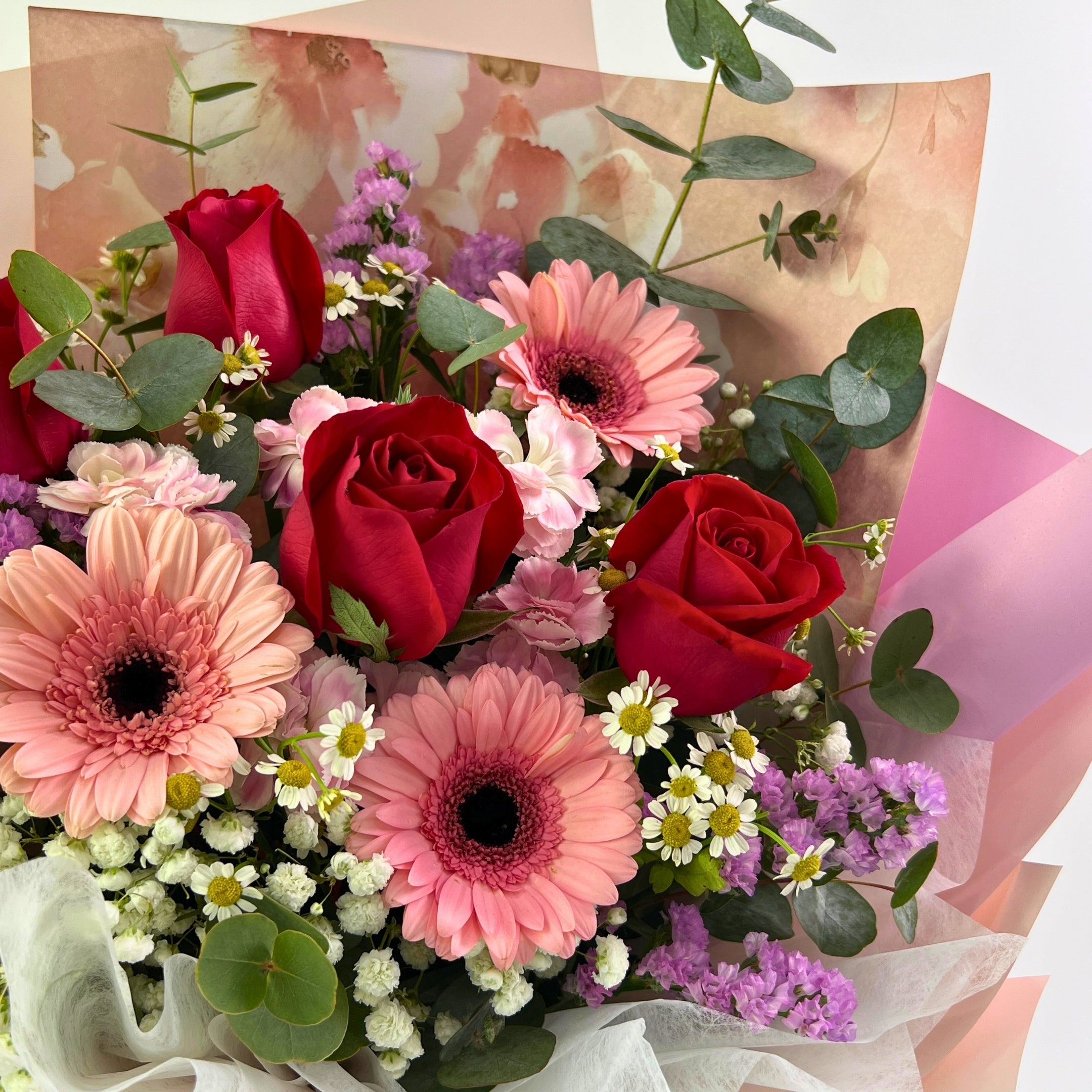 Bouquet of flowers with red roses, pink gerberas, and greenery on a soft pink background.