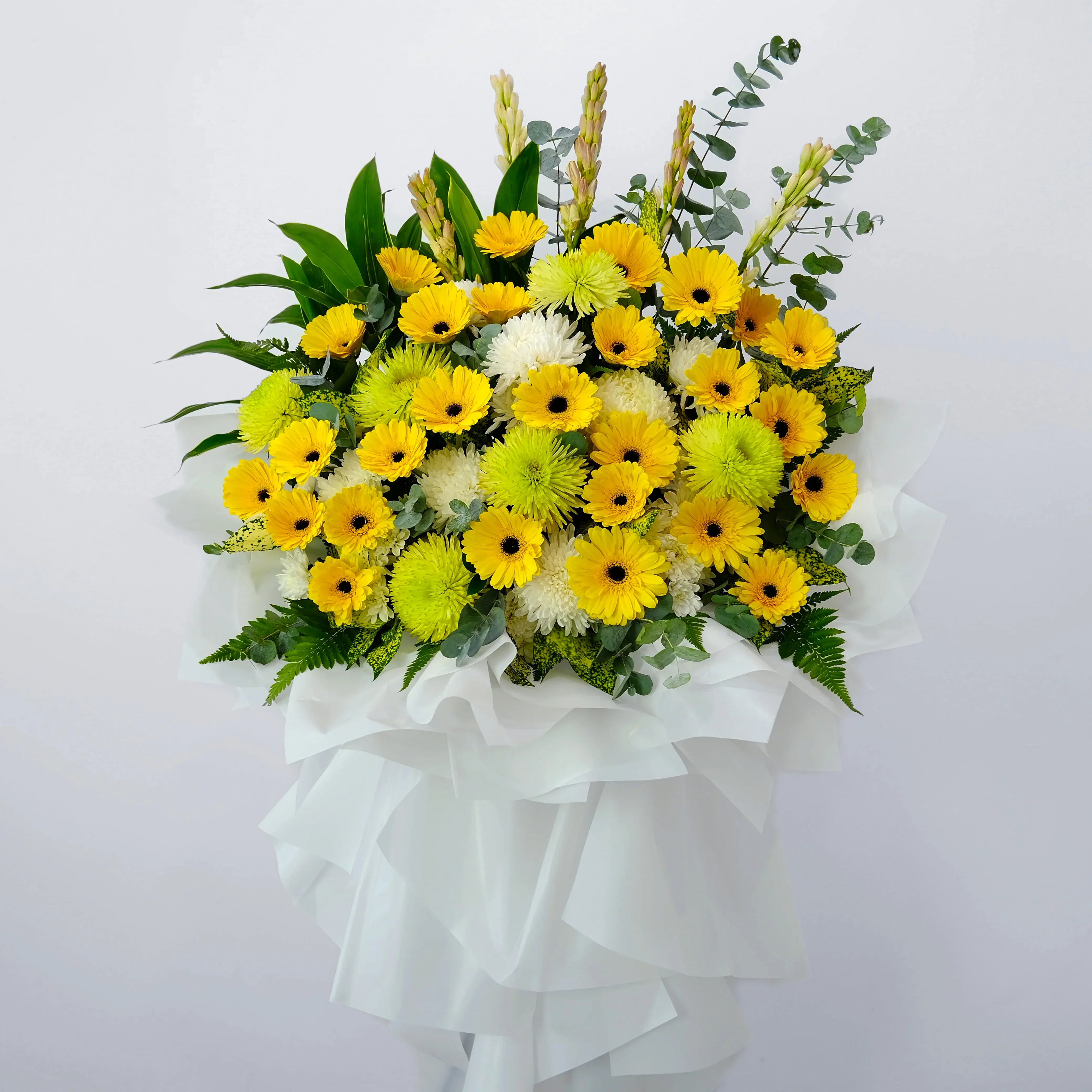 Yellow gerbera daisies and white chrysanthemums arrangement with green leaves, wrapped in white.