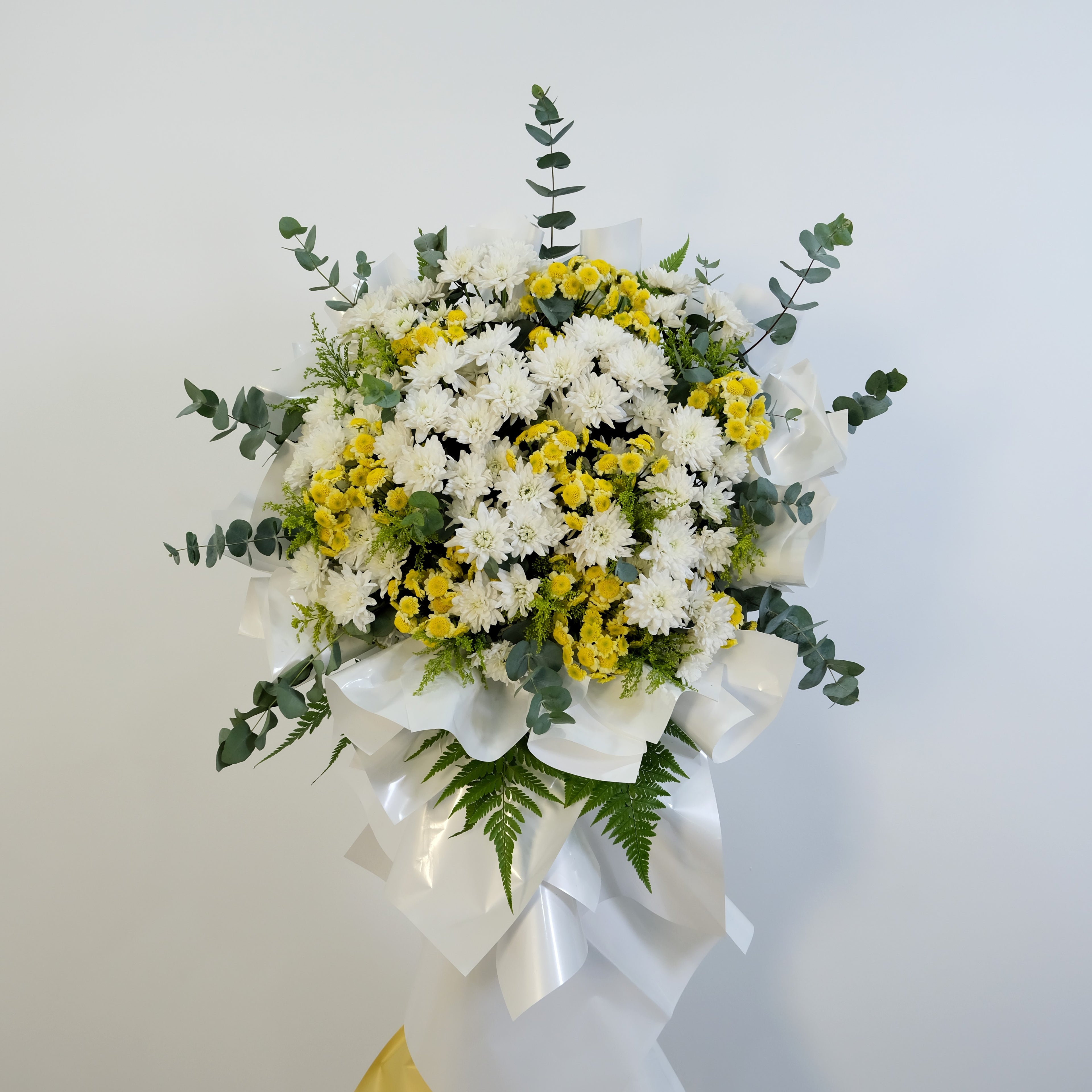 condolence flower stand of white and yellow flowers with greenery on a light gray background