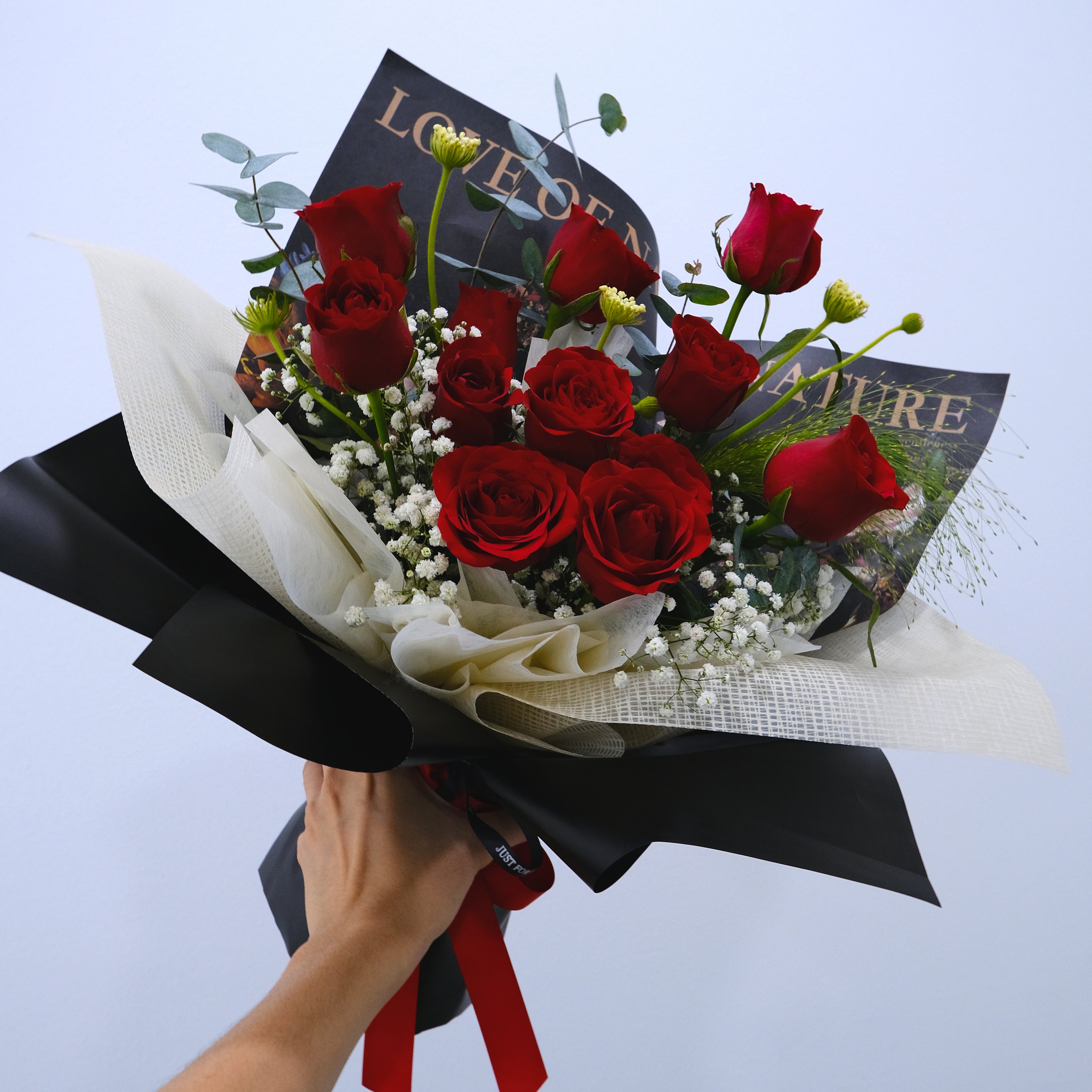 Bouquet of red roses wrapped in black paper with a hand holding it against a light blue background.