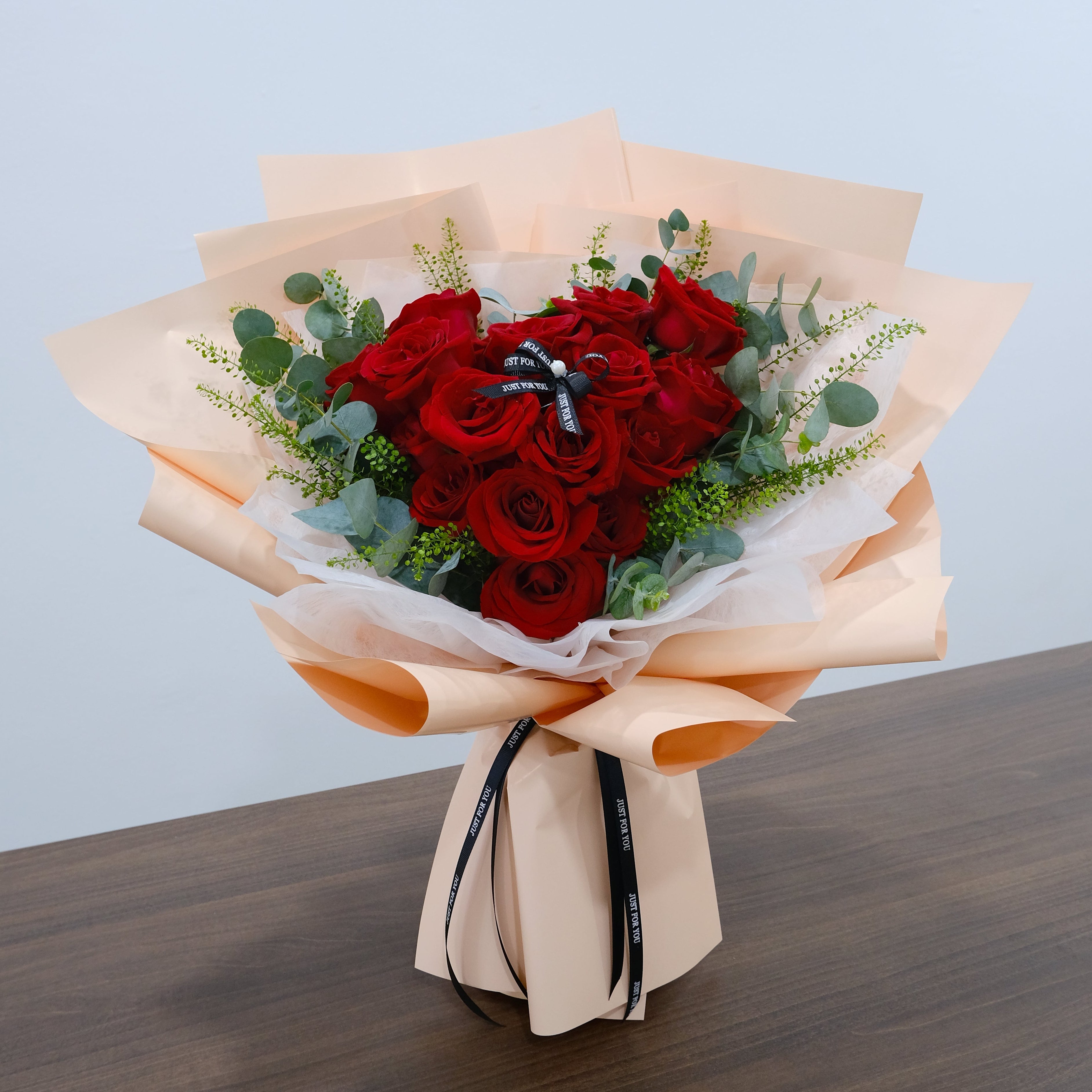 Bouquet of red roses with greenery wrapped in brown paper on a wooden surface.