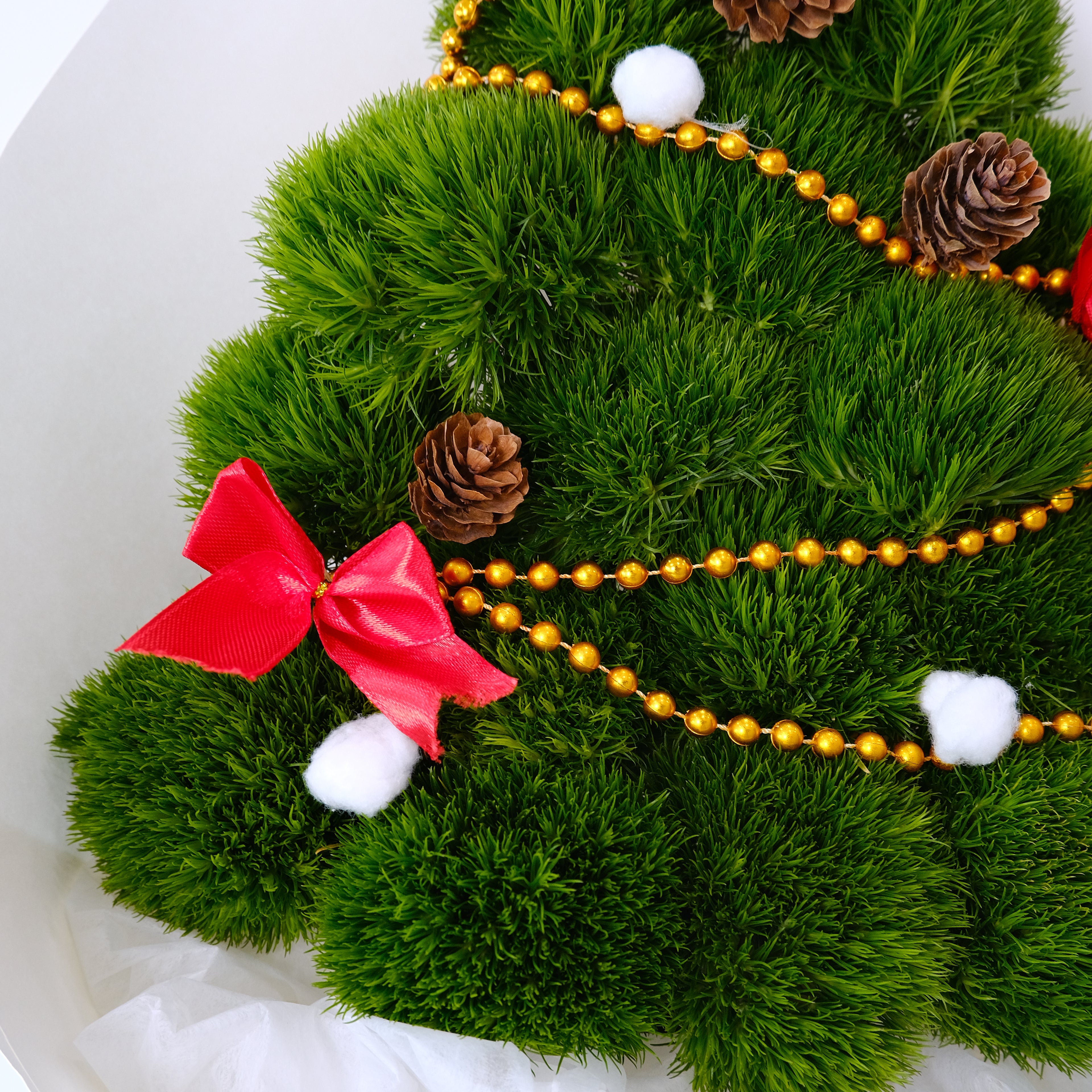 Decorative green wreath with a red bow, gold beads, pinecones, and white pom-poms on a white background.