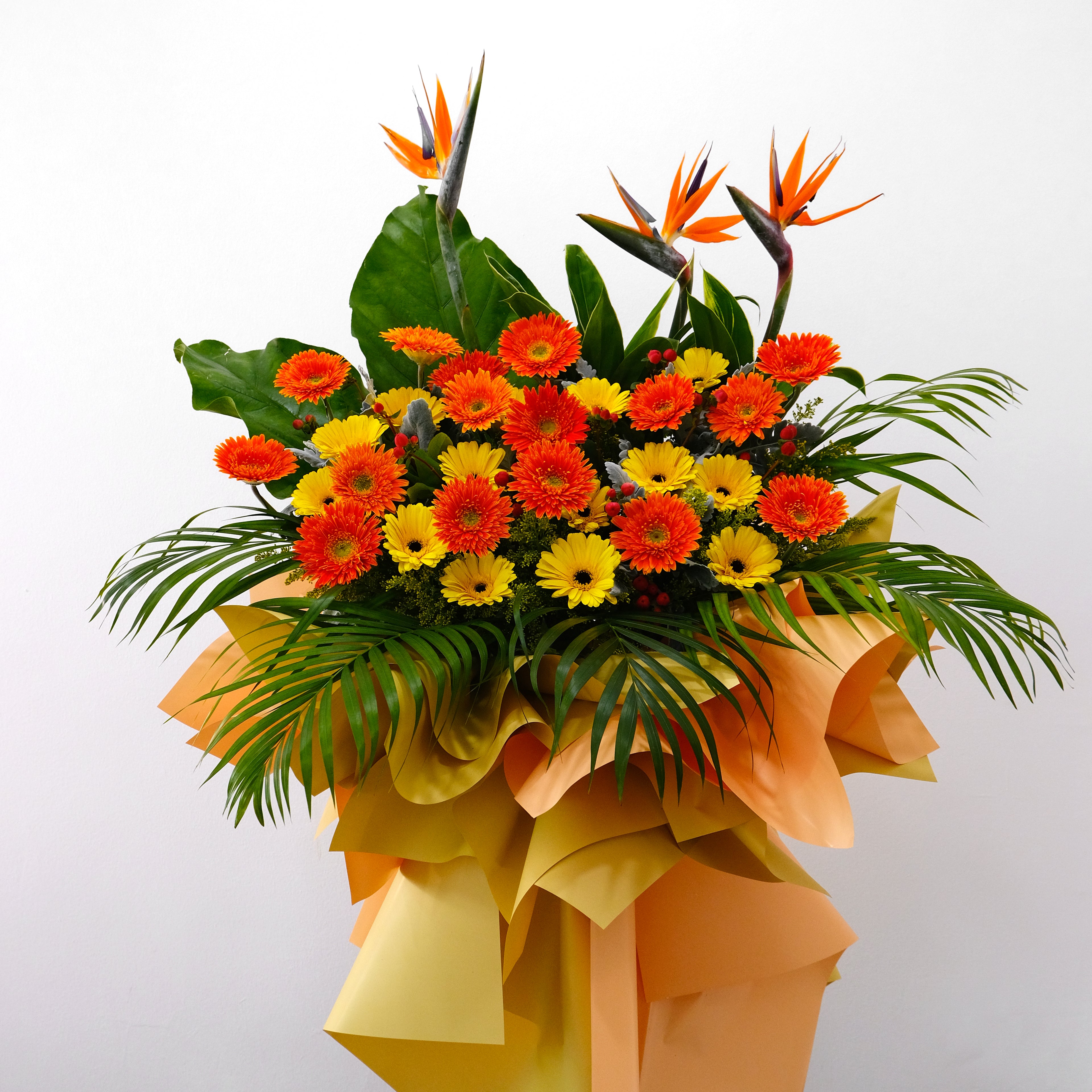Grand opening flower stand featuring Bird of Paradise, orange gerberas, and palm leaves in a gold and orange designer wrap for luxury delivery in KL.